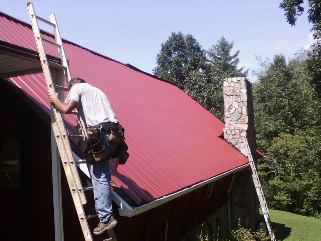 Roofer on a ladder beside a red metal roof and stone chimney — Soffit & Fascia Repair by Roofing Brothers in Monkton